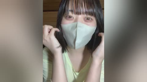 Video of an Asian woman with straight black hair, wearing a light green surgical mask, light yellow shirt, and holding her face with both hands. Background blurred, showing wooden panels.