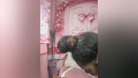 Media: Video of a young woman with dark hair in a bun, wearing a light-colored top, bending over in a pink room with floral wallpaper and a decorative bow on a white door.