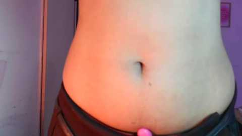 A close-up video of a person's lower torso, featuring a navel piercing and a hint of black underwear, set against a purple wall.