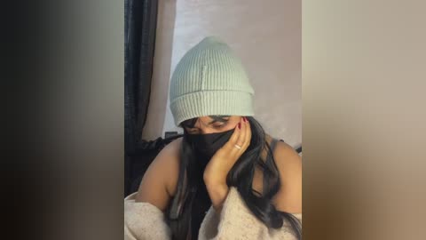 Video of a woman with long black hair, wearing a light blue knit beanie, black face mask, and a beige sweater, sitting indoors with a neutral background.