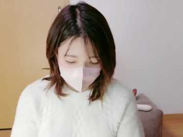 Video of an Asian woman with shoulder-length brown hair, wearing a white mask, sitting indoors with a wooden floor and beige wall. She has a calm expression, and her hands are visible, holding a white object.