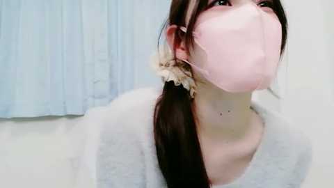 Video of a young Asian woman with fair skin, brown hair tied in a low ponytail, wearing a pink surgical mask, light gray sweater, and beige scrunchie. Background features light blue curtains and a white floor.