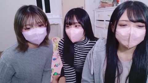 Video of three young Asian women wearing light pink surgical masks, seated indoors. They have long black hair and wear casual clothes, including striped shirts and a gray jacket. Background shows white furniture and a window.