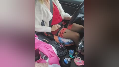 Video of a woman in a red dress, beige cardigan, and black stockings, sitting in a car with a pink car seat, steering wheel, and visible child seat in the background.