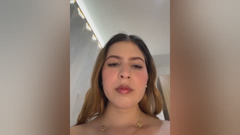 Video of a young woman with fair skin, long dark hair, and light makeup, wearing a white top and a necklace, standing in a well-lit room with a modern ceiling light.