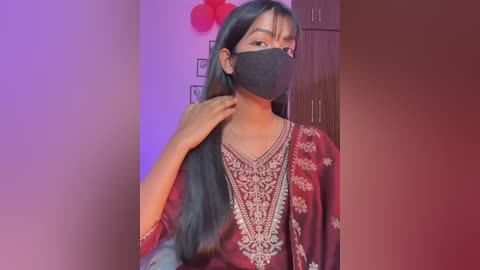 Video of a South Asian woman with long black hair, wearing a maroon sari with intricate embroidery, black face mask, and red flower behind her head.