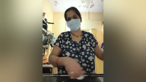 Media: Video of a woman with dark hair and medium skin tone, wearing a blue floral shirt, blue face mask, and a red necklace, cleaning a black countertop in a yellow-painted room.