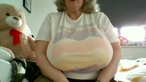 A video of a woman with large breasts, wearing a white t-shirt with a pastel rainbow design. She sits on a bed with a teddy bear, in a bright, softly lit room.
