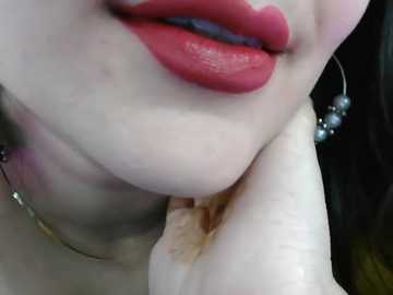 A close-up video of a light-skinned woman's face with full, glossy red lips, holding her chin, wearing silver hoop earrings. Background is dark, focusing on her lips and hand.