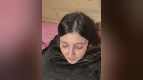 Video of a young woman with long dark hair, wearing a black top, lying on a pink bedspread, eyes closed, against a wooden headboard.