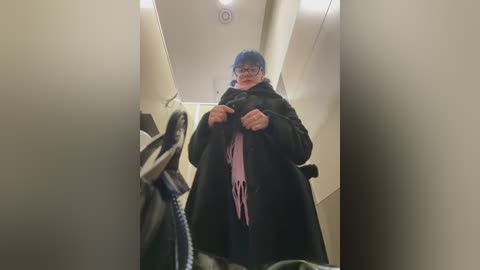 A video of a woman in a store, wearing glasses, blue hat, and black coat, holding a pink scarf, surrounded by beige walls and ceiling lights.
