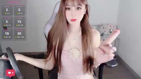 Video of a young East Asian woman with long, straight brown hair, wearing a light pink spaghetti strap top, sitting in a gaming chair, holding a controller, in a minimalist room with a white wall and a calendar overlay.