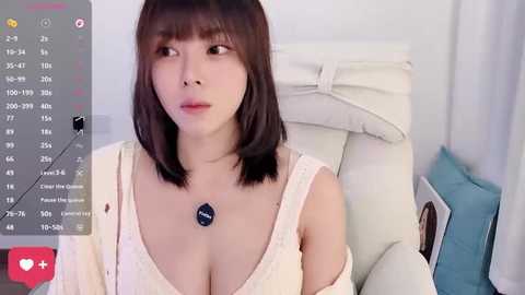 Video of a young Asian woman with straight brown hair, wearing a low-cut white lace top, sitting in a white chair. The background shows a bed with white sheets and blue pillows.