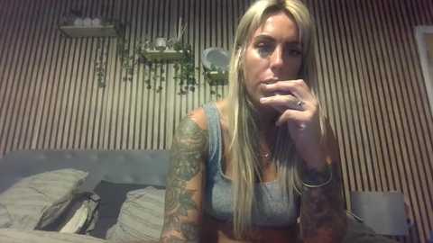 Video of a tattooed, blonde woman in a gray tank top, sitting on a bed with striped wallpaper in the background.