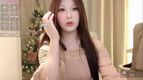 Video of a young Asian woman with long brown hair, wearing a beige lace dress, holding a phone, sitting in a room with a Christmas tree.