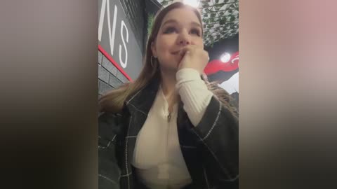 A video of a smiling young woman with fair skin and long blonde hair, wearing a black plaid coat over a white top, standing in front of a brick wall with greenery and a red canopy in the background.