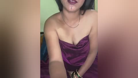 Video of a woman with medium brown skin and dark hair, wearing a purple satin dress, with a gold bracelet, and a small nose stud.