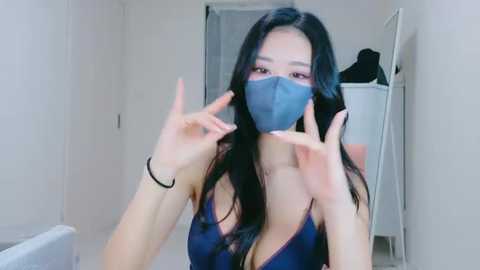 A video of an Asian woman with long black hair, wearing a blue face mask, dark blue lingerie, and black bracelets, standing in a narrow hallway.
