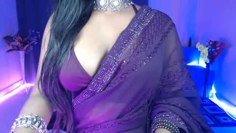 Video of a woman with long black hair, wearing a deep purple, intricately embroidered sari with a plunging neckline, adorned with silver jewelry. Background features blue lighting and decorative floral arrangements.