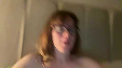 A blurred video of a Caucasian woman with shoulder-length brown hair and glasses, likely in a bathroom, wearing a tank top. The background is out of focus, featuring a beige wall and a partially visible sink.