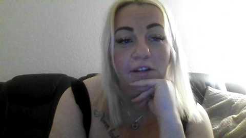 A video of a middle-aged woman with straight, shoulder-length blonde hair, fair skin, and a slightly pensive expression. She wears a black top, with a pendant necklace visible. Background features a dark fabric sofa and light wall.