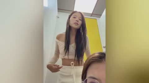 Video of a slim, young Asian woman with long black hair in a white off-shoulder crop top and high-waisted pants, standing in a brightly lit, white-walled bathroom with a yellow door.