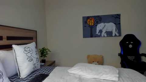 Video of a minimalist bedroom with a beige wall, a wooden headboard, white bedding, and a black gaming chair. A framed artwork of a white elephant and a colorful tree hangs above the headboard.