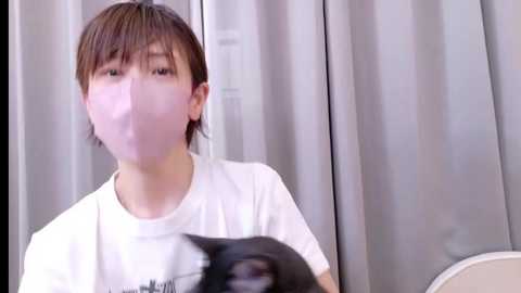 Video of a young Asian boy with short brown hair, wearing a white t-shirt, and a surgical mask. He holds a black cat in his hands. Background features gray curtains.