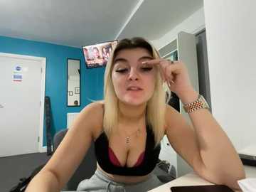 Video of a young, blonde-haired woman with fair skin, wearing a black sports bra, gray sweatpants, and a gold bracelet. She sits in a modern office with blue walls, a white door, and a TV displaying a video.