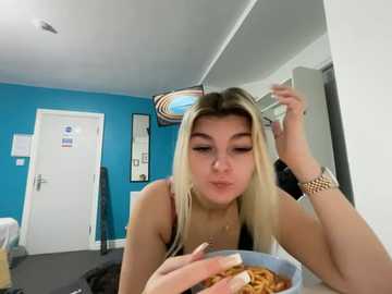 Video of a young woman with long, blonde hair, wearing a black top, eating noodles in a modern room with blue walls, white door, and a mirror.