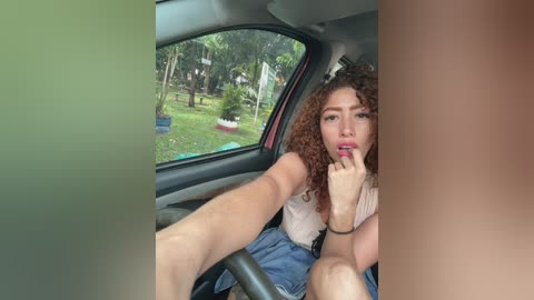 Video of a young woman with curly hair, light skin, and full lips, wearing a white top and blue shorts, sitting in a car, looking out the window at a green park with trees and a red bench.