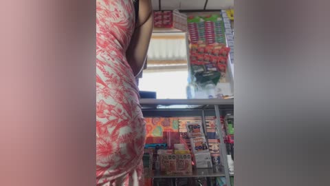 A video captures a woman's torso in a red floral dress, reaching for items on a supermarket shelf. The background showcases various colorful packaged goods, including snacks and drinks. The scene is well-lit, highlighting the vibrant colors.