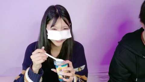 Video of an Asian woman with long black hair wearing a surgical mask, eating ice cream from a cup with a spoon. She's dressed in a black shirt. The background is a light purple gradient.
