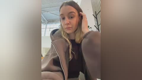 Media: Video of a young Caucasian woman with straight, blonde hair, wearing a light brown jacket and a dark top, looking introspective. Background features a modern indoor setting with large windows and metallic ceiling beams.