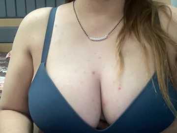 A close-up video of a light-skinned woman with large breasts wearing a blue bra. She has a silver necklace, and her long, brown hair is partially visible. The background shows a bed with patterned sheets.