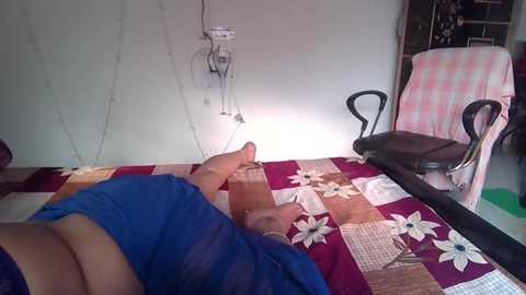 Media: Video of a person lying on a bed with a patchwork quilt, wearing a blue shirt, legs extended, in a modest, sparsely furnished room with a chair and a partially visible wall-mounted medical device.