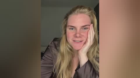 Video of a smiling Caucasian woman with long blonde hair, wearing a dark blouse, resting her chin on her hand, set against a blurred indoor background.