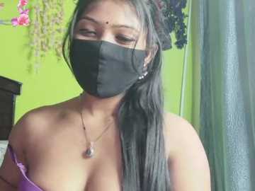 Video of an Asian woman with long black hair, wearing a black face mask, a purple strapless top, and a necklace. She stands indoors with green walls and a floral decoration in the background.