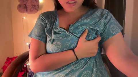 Video of a woman with medium skin tone, wearing a turquoise hospital gown with white floral patterns, holding her breast, in a dimly lit room with string lights, a plush toy, and a red bag.