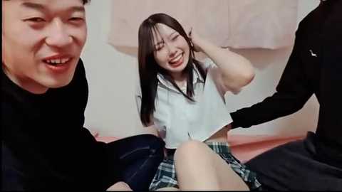 Media: A video captures a smiling Asian woman in a white blouse and plaid skirt, seated on a pink bed, surrounded by two men in black clothing.
