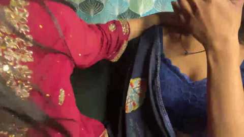 Video of two women, one in a vibrant pink sari with intricate golden embroidery, the other in a blue lace top, holding a teal cloth.