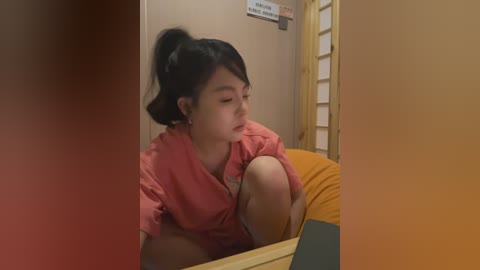 A video of a young Asian woman with black hair tied in a high ponytail, wearing a pink t-shirt, sitting on a wooden bench in a dimly lit, beige-walled room.