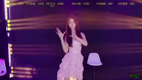 Video of a young Asian woman with long red hair singing into a microphone, dressed in a pink frilled dress, standing against a dark backdrop with white lights.