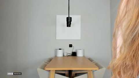 Video of a minimalist dining room with a light wooden table, white chairs, a wall-mounted light, and a single person's long blonde hair in the foreground.