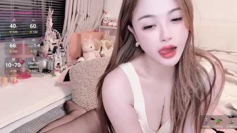 A video of a young Asian woman with long, straight brown hair and fair skin, wearing a white lace bra, sitting on a bed with teddy bears, surrounded by a cozy room.