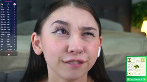 Media: Video of a young woman with straight black hair, wearing a nose ring, white AirPods, and a light pink lip gloss. Background shows a gray headboard and a green plant.