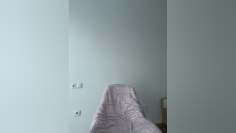 Video of a minimalist room with a light gray wall, a white outlet, and a plaid-patterned pink blanket draped over a chair, set against a simple, serene background.