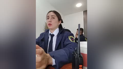 Video of a young woman with long brown hair in a navy blue police uniform, gesturing with her hands, in a room with white walls and a wooden table, blurred background.