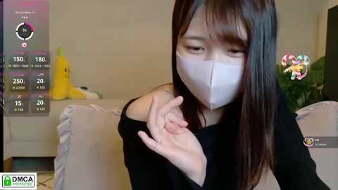 Video of an East Asian woman with long black hair wearing a white mask, black top, and sitting indoors. Background includes a gray couch, plush toys, and a live streaming interface.