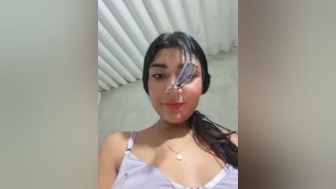 A video of a young Latina woman with medium-dark skin and long dark hair in a ponytail. She wears a white spaghetti-strap tank top and a gold necklace. Her face is partially obscured by a butterfly, creating a playful, whimsical effect.